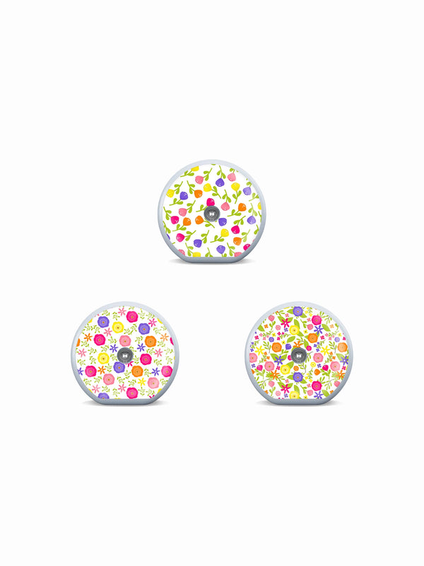 Three Accu-Chek SmartGuide sensor stickers with colorful buds, flowers and garden-inspired patterns on white.